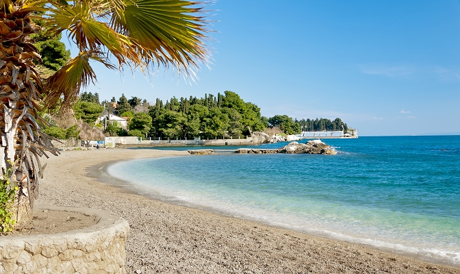 Na slici je pješčano-šljunkovita plaža uz obalu mora. U prvom planu nalazi se palma, dok su uz obalu vidljivi tirkizno more, stijene i uređena šetnica. U pozadini se nalaze drveće, nekoliko objekata i vedro plavo nebo.