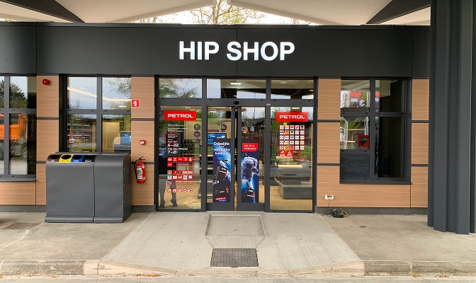 Na slici je ulaz u Hip Shop na Petrol prodajnom mjestu. Iznad staklenog ulaza nalazi se veliki natpis „HIP SHOP“. Pored ulaza postavljene su kante za odvojeno prikupljanje otpada i protivpožarni aparat. Na ulaznim vratima i izlozima vidljive su različite informativne i promotivne naljepnice.