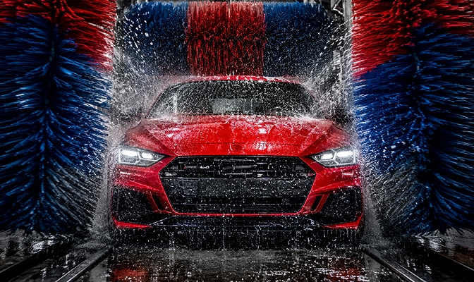 Na slici je crveni automobil u automatskoj autopraonici. Vozilo se nalazi između velikih rotirajućih četki plave i crvene boje, dok voda prska po automobilu tokom procesa pranja.