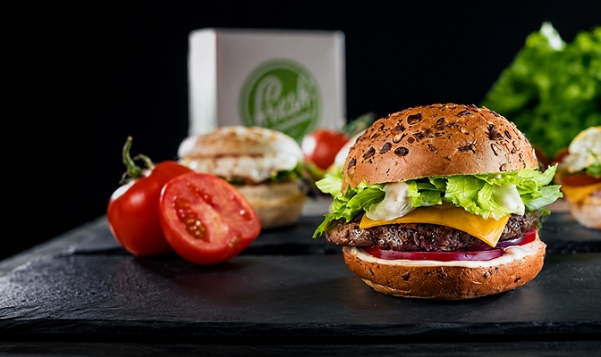 Na slici je hamburger sa zelenom salatom, sirom, mesom i crvenim lukom, postavljen na tamnoj podlozi. Pored hamburgera nalaze se svježi paradajzi, dok su u pozadini vidljivi dodatni hamburgeri i zelena salata.
