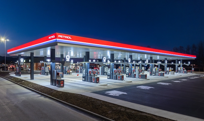 Na slici je Petrol benzinska stanica sa više točionih mjesta za gorivo. Stanica je osvijetljena i natkrivena velikom nadstrešnicom sa Petrol oznakama. U prvom planu vide se točilice za gorivo, saobraćajne oznake i pristupne saobraćajnice.