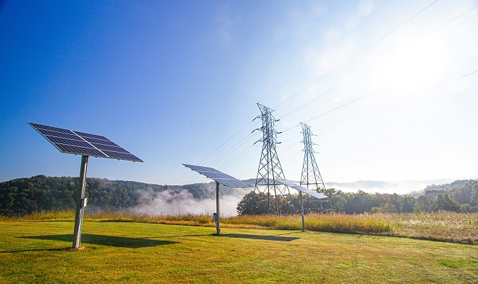 Na slici su solarni paneli postavljeni na travnatoj površini. U pozadini se nalaze dalekovodi i stubovi za prijenos električne energije, dok su iza njih vidljivi drveće, brda i vedro nebo.