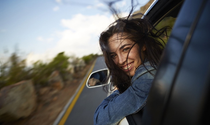 Na slici je osoba nagnuta kroz otvoren prozor automobila. U pozadini je vidljiva cesta koja prolazi kroz prirodni krajolik, dok se uz vozilo nalaze dijelovi ceste i vegetacije.